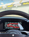 A selfie-style view from inside a car, showing a large black steering wheel and a digital instrument display with a red and black graphic. A sign in the background reads Haltech, and a text overlay says 'Today's office @santapodraceway'.