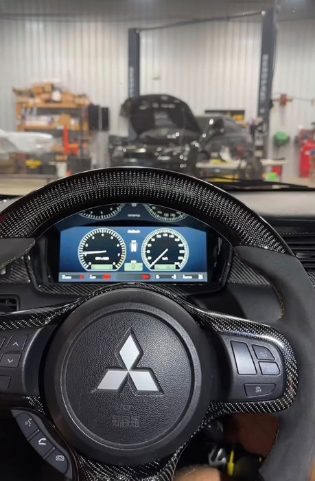 A close-up of a Mitsubishi steering wheel with a carbon fiber pattern and the brand's three-diamond logo, positioned in front of a car's digital instrument cluster. The background shows a blurred auto repair shop with cars and equipment.