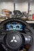 A close-up of a Mitsubishi steering wheel with a carbon fiber pattern and the brand's three-diamond logo, positioned in front of a car's digital instrument cluster. The background shows a blurred auto repair shop with cars and equipment.