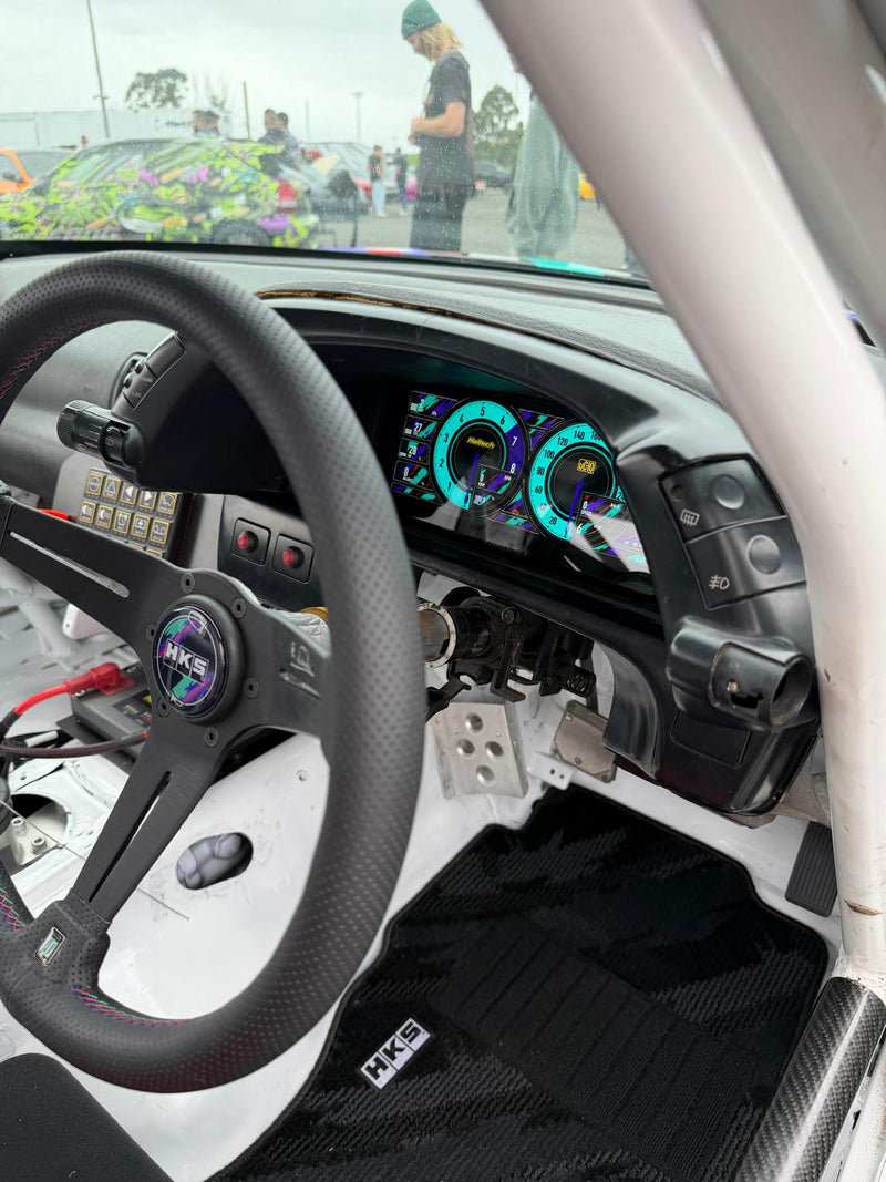 A close-up view of a custom HKS racing cockpit interior, featuring a black HKS steering wheel, a roll cage, a white interior base, and a custom digital instrument cluster with glowing teal gauges. The car is photographed from the passenger side inside the vehicle, with a race track and people visible through the windshield in the background.