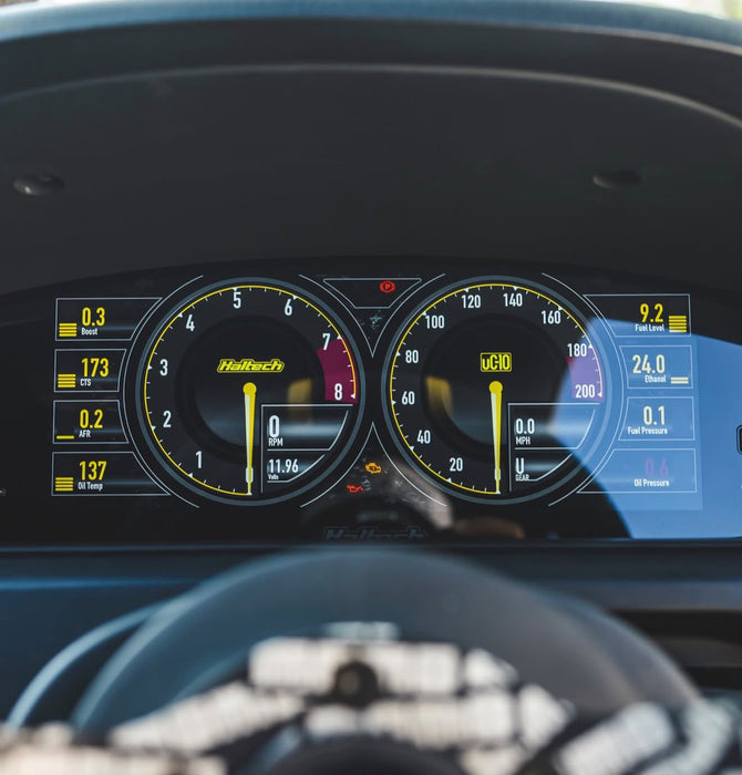 Close-up of a car's digital instrument cluster with two large analog-style gauges and bright yellow markings, mounted inside a vehicle dashboard above a blurred steering wheel.