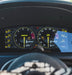 Close-up of a car's digital instrument cluster with two large analog-style gauges and bright yellow markings, mounted inside a vehicle dashboard above a blurred steering wheel.