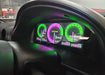 A close-up of a car instrument cluster with a custom digital display showing bright green and purple gauge graphics inside a black dashboard.